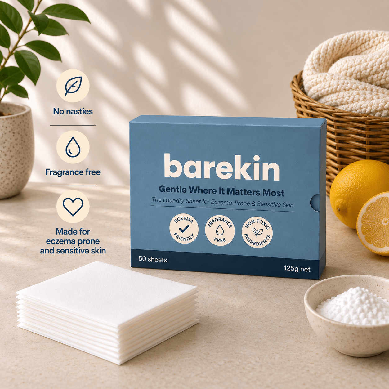 A box of barekin laundry sheets stands on a beige surface with a stack of white sheets in front and a bowl of white granules to the right. A basket of folded towels, lemons, and a potted plant appear in the background, along with marketing callouts reading 'No nasties,' 'Fragrance free,' and 'Made for eczema prone and sensitive skin.